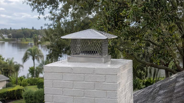 A stainless steel mesh chimney cap covered with raindrops on a white-painted brick chimney.